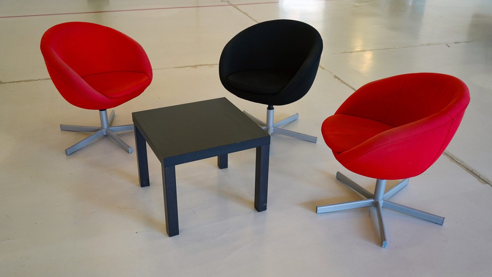 Two red swivel chairs and one black swivel chair arranged around a small table on a bare concrete floor