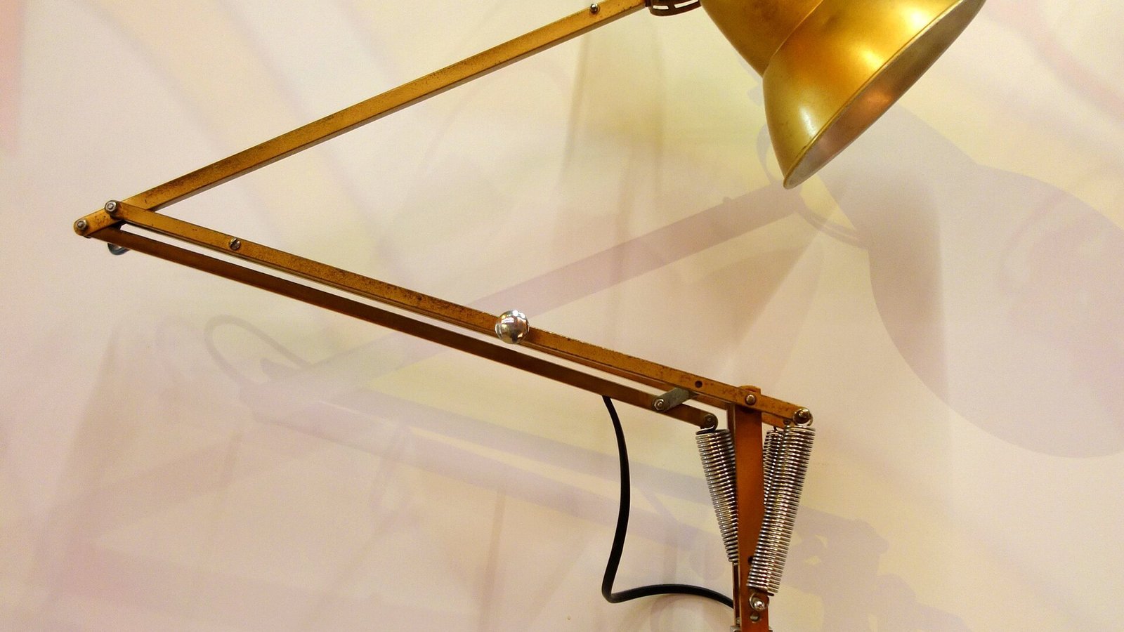 Close-up of a brass gooseneck desk lamp's spring-balanced swing arm and shade against a plain wall