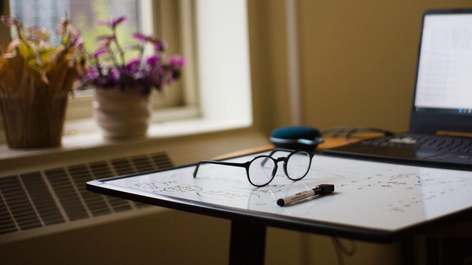 A small whiteboard lying flat on a desk with glasses and a dry erase marker on it, a laptop behind and potted flowers by the window