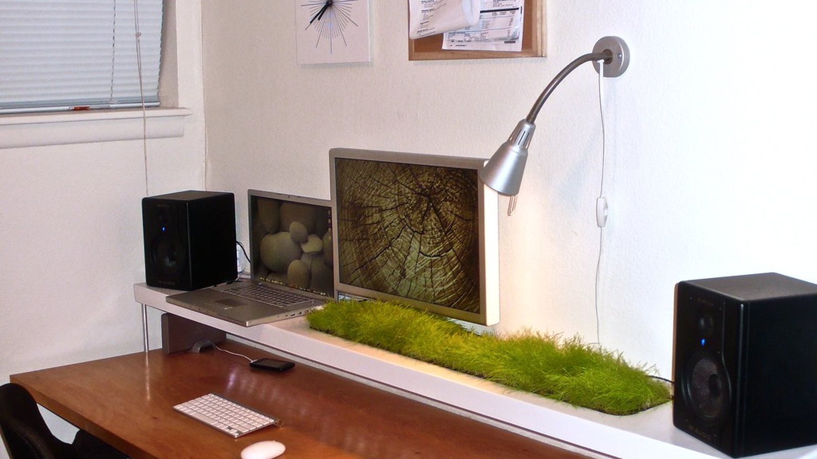 A compact desk setup with a laptop, small speakers, and a wall-mounted lamp against a plain white wall