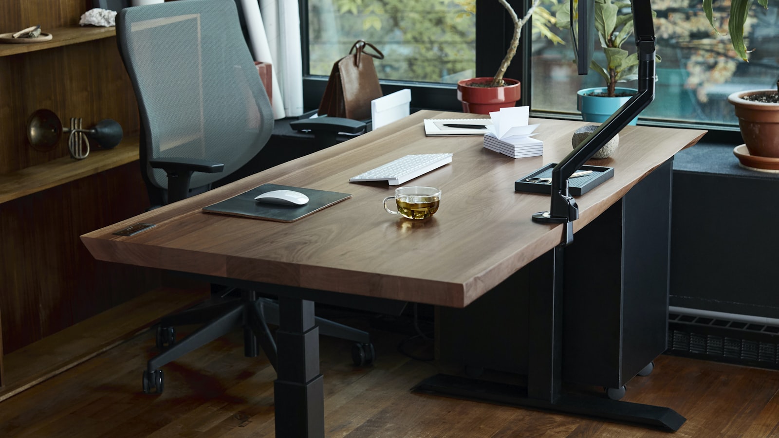 A height-adjustable wooden desk with a monitor arm, keyboard, and mouse pad, in a bright room