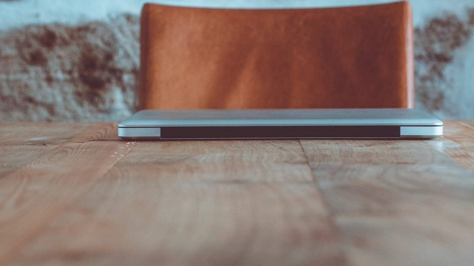 A closed silver laptop on a wooden desk with a leather chair back blurred in the background