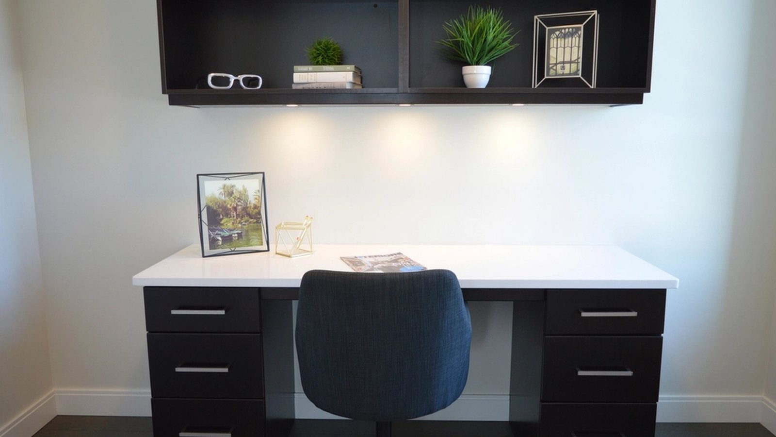 A desk lit from above by fixtures mounted under the shelf over it, throwing light down onto the surface