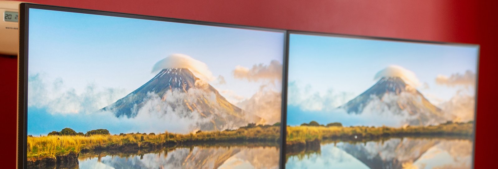 Two monitors mounted side by side on a dual arm, both showing the same volcano wallpaper that repeats instead of continuing across the bezel gap