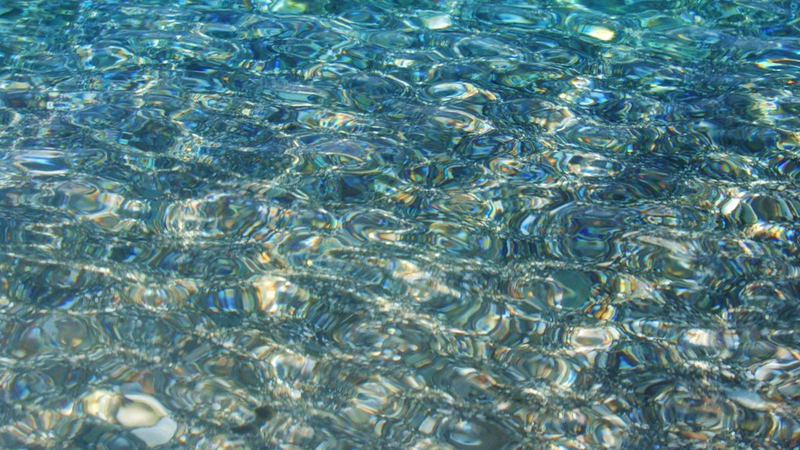 Extreme close-up of clear, sunlit turquoise sea water rippling over pale sand and pebbles