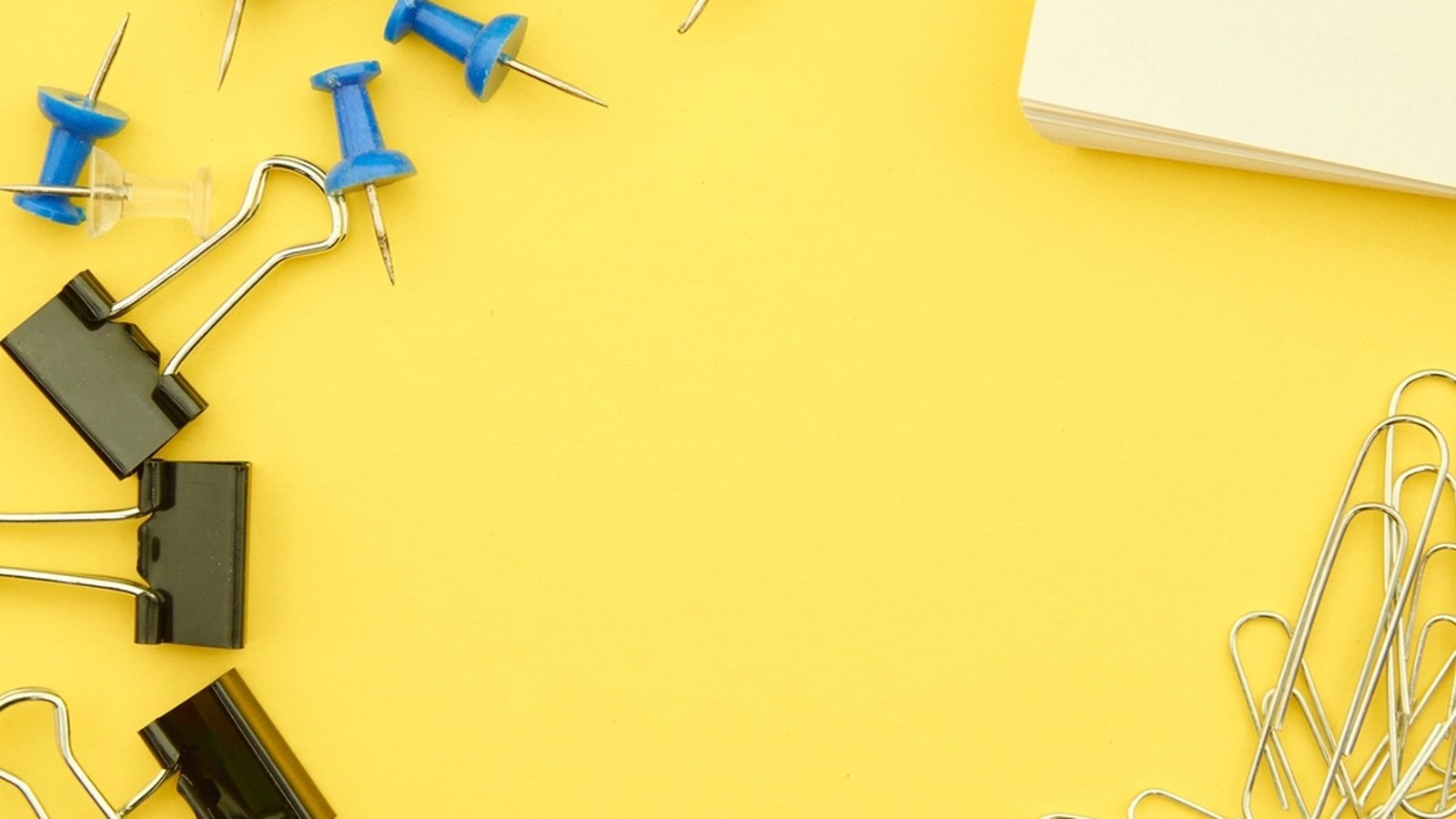 Push pins, binder clips and paper clips scattered on a yellow desk background