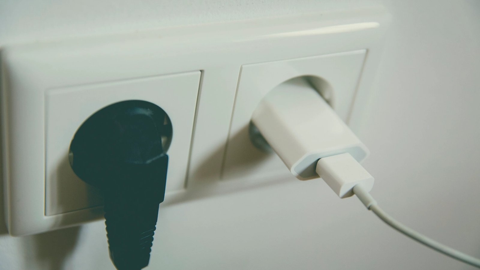 Close-up of a wall outlet with a black plug and a white charging cable plugged in side by side