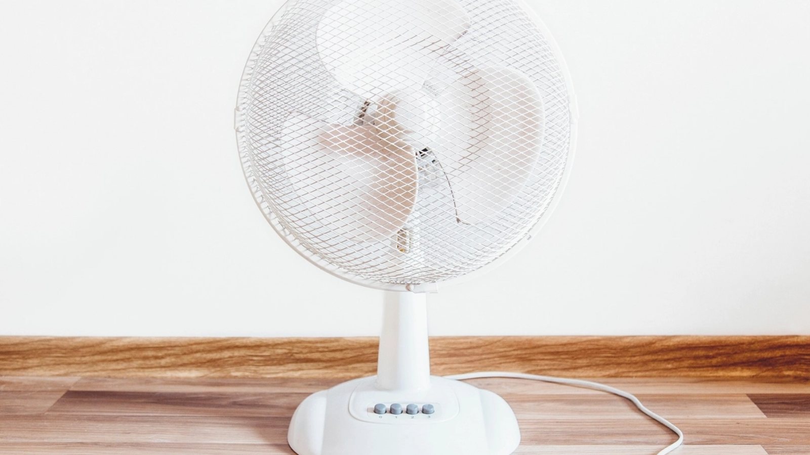 A white table fan with visible speed buttons and a trailing cord, standing on a wooden surface against a plain wall